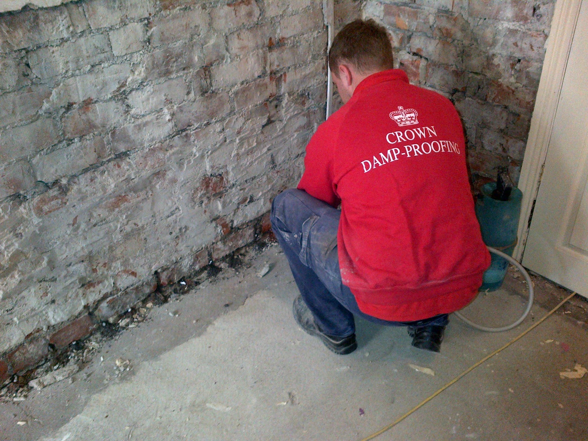 A Crown Damp-Proofing operative in a red branded jacket injecting a new damp course along a stripped brick wall